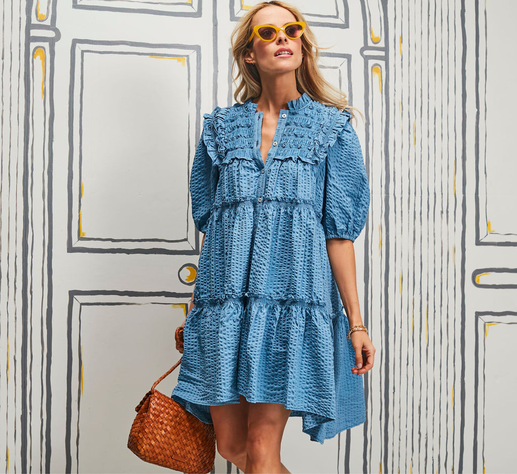 Woman in a classic, light-blue dotted babydoll dress with ruffled smocked bodice, carrying a woven tote. Effortless, high-end style with fun accessories.