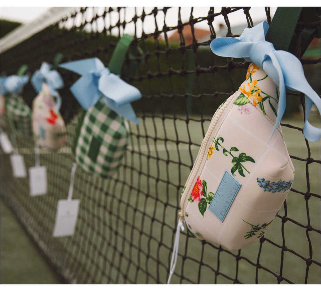 Decorative pouches in classic patterns, tied to a tennis net with powder-blue bows, showcasing Tuckernuck's effortless, fun, high-end style.
