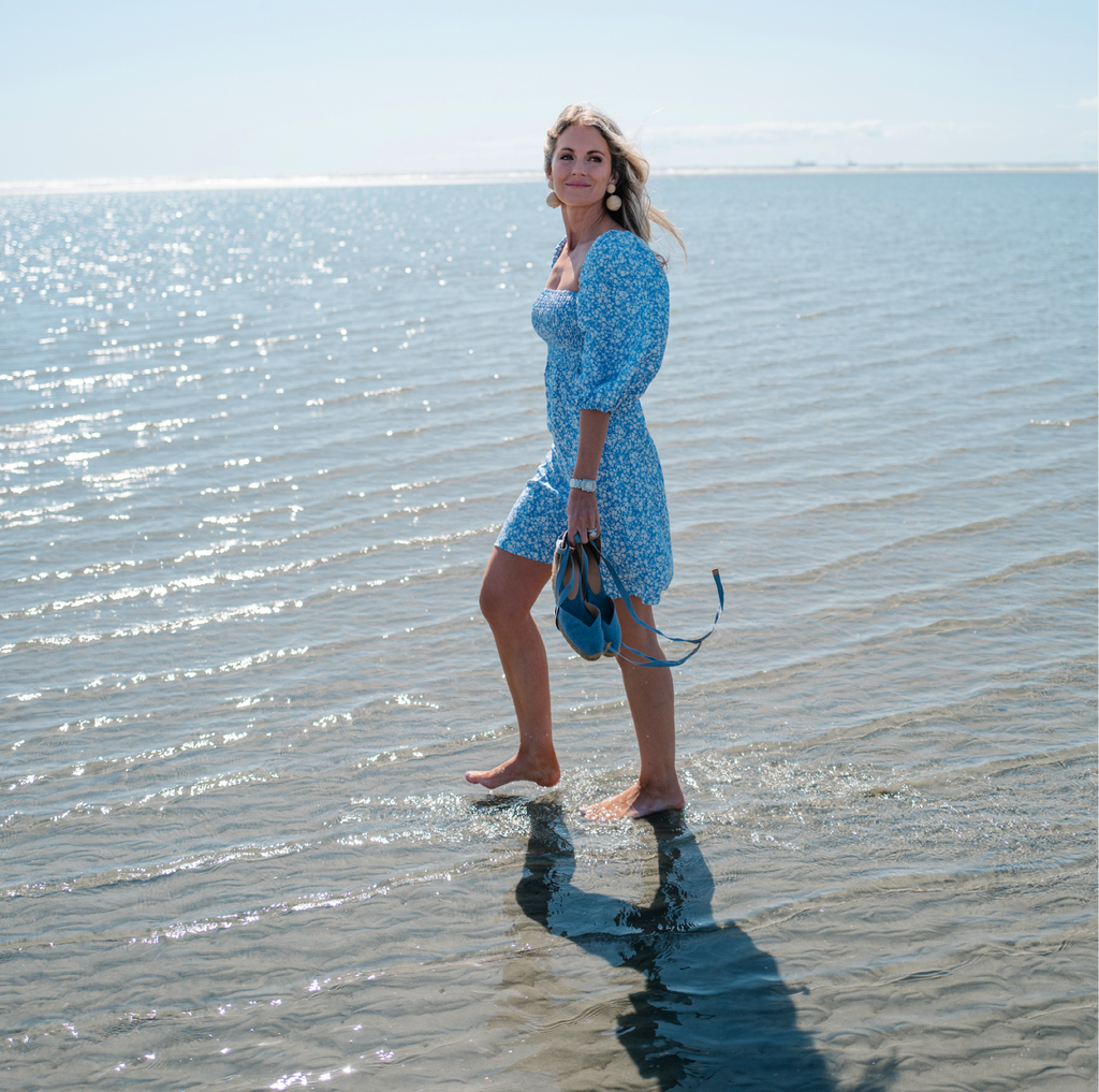 Woman in a blue floral dress walks barefoot along a sunlit beach, carrying classic blue wedges, embodying effortless, high-end style for fun seaside moments.