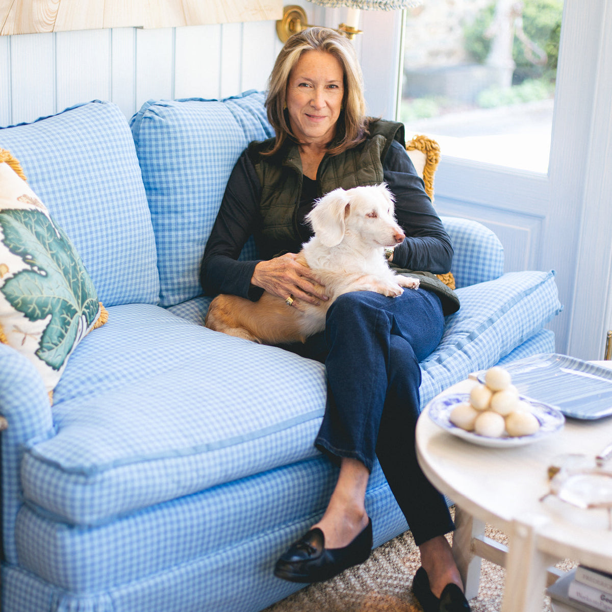 A woman in a classic, light-blue gingham sofa with a white dog; round coffee table nearby, creating an effortless, fun, high-end living room vibe.