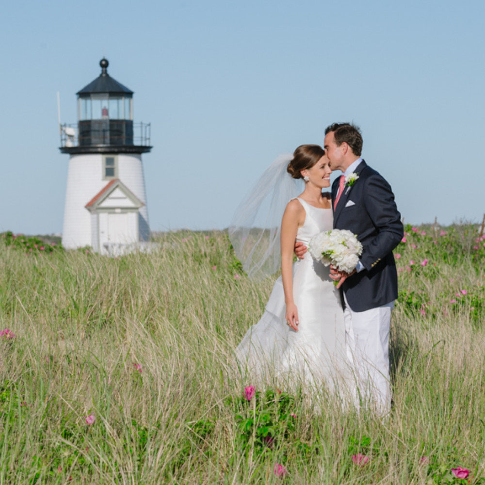 Bride and groom in classic, effortless wedding attire share a tender moment in a field with pink wildflowers, near a lighthouse, embodying high-end, fun elegance.