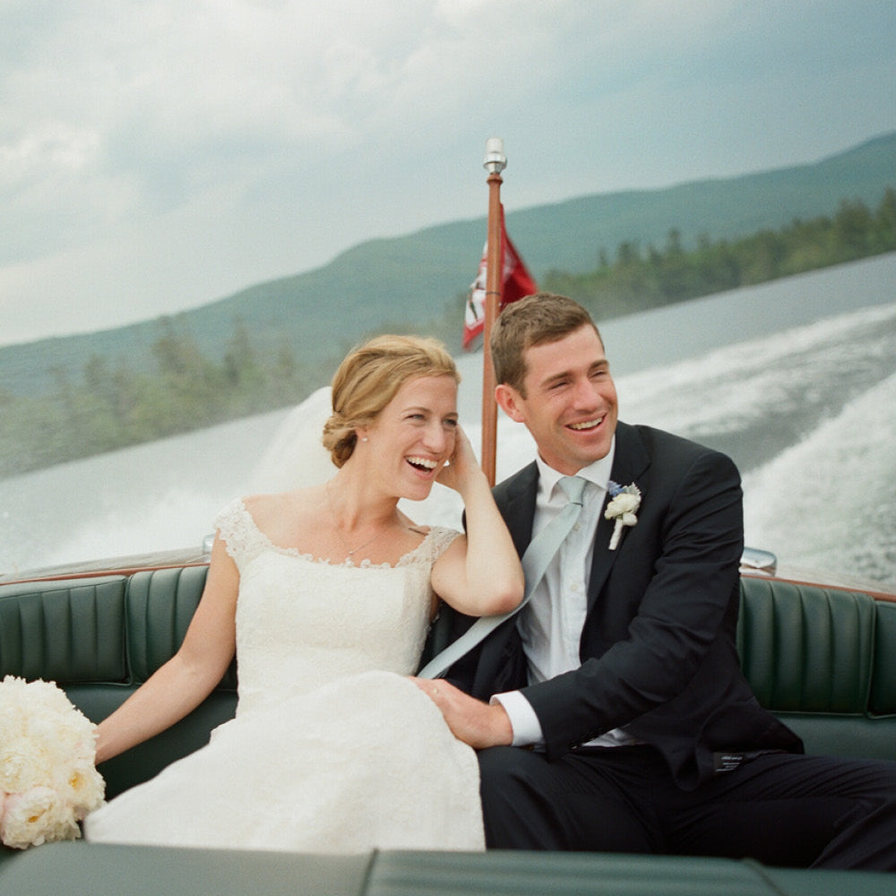 Newlywed couple enjoying an effortless, fun ride in a motorboat; bride in classic lace gown, groom in high-end suit, both smiling amidst scenic backdrop.