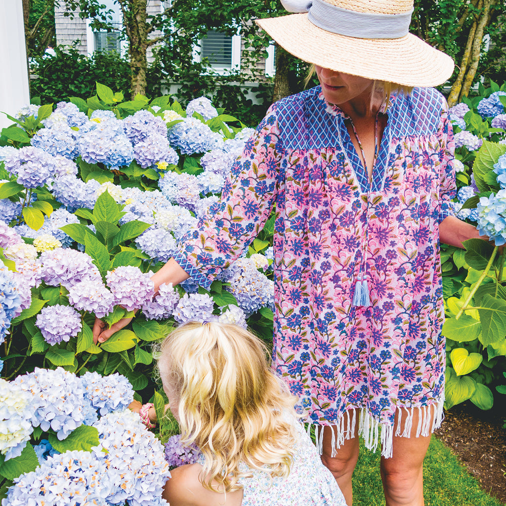 A woman in a straw hat and floral tunic, with a child, enjoy hydrangeas in a garden, embodying classic, effortless, and fun high-end style.