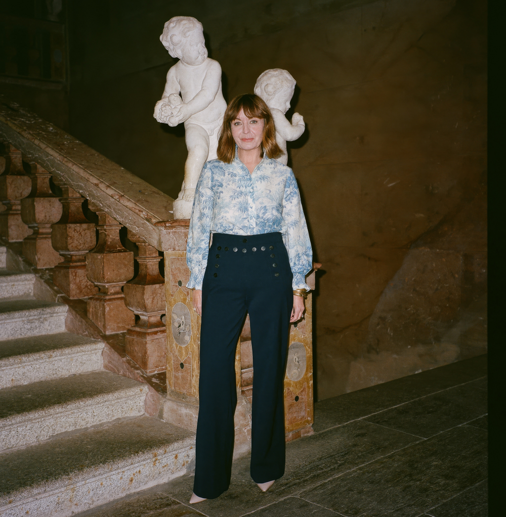 Woman in classic, floral blouse and high-waisted navy trousers stands by ornate staircase; embodies effortless, high-end style with fun, historic ambiance.