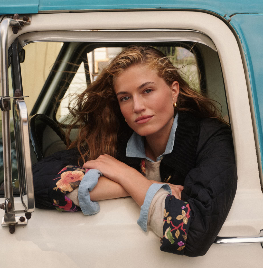 Young woman in classic, high-end quilted jacket with floral lining, leaning out a vintage pickup, embodying effortless, fun style.
