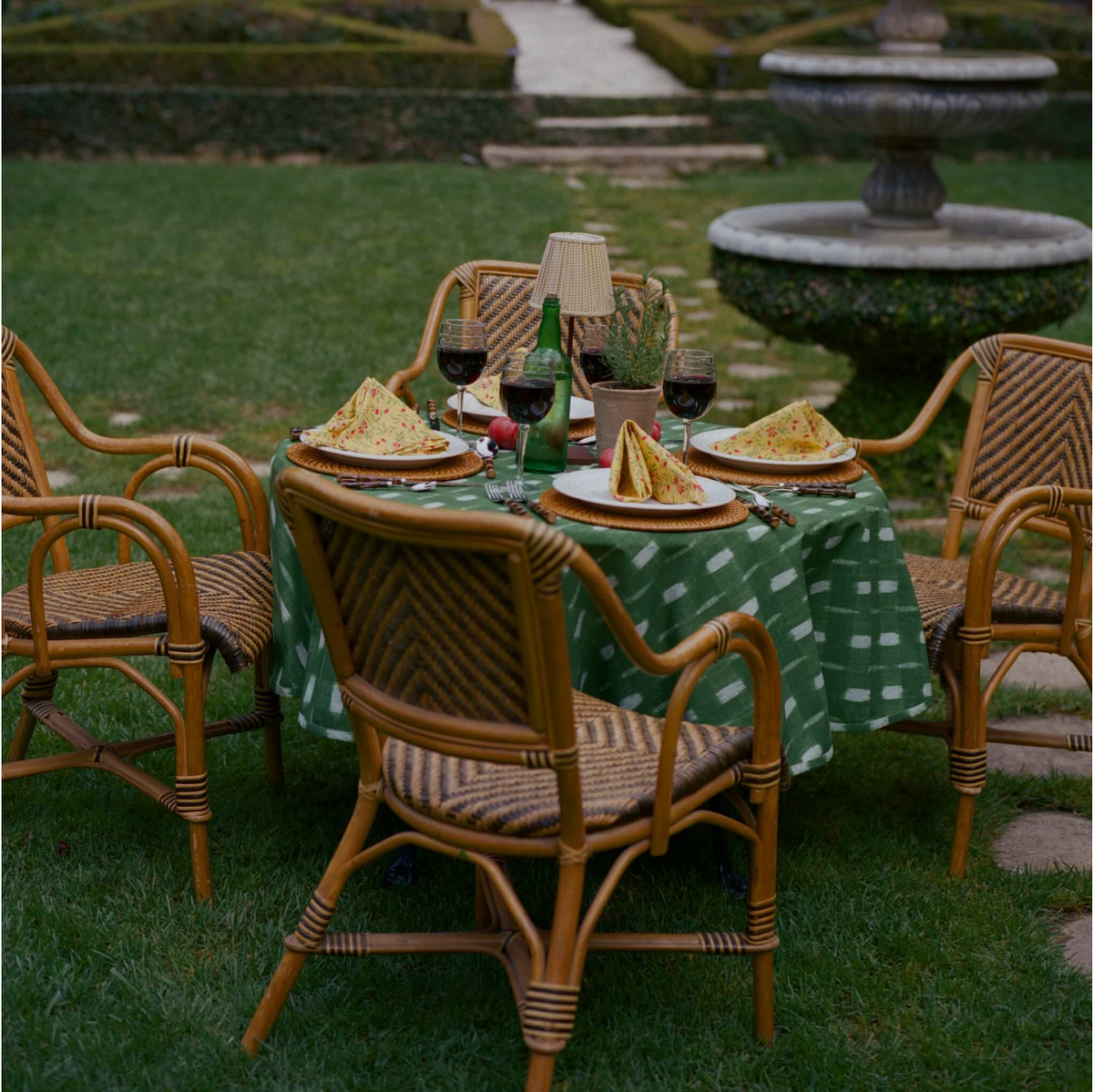 Classic al fresco dining with an effortless setup: green tablecloth, wicker chairs, wine, and floral napkins—perfect for a fun, high-end garden experience.