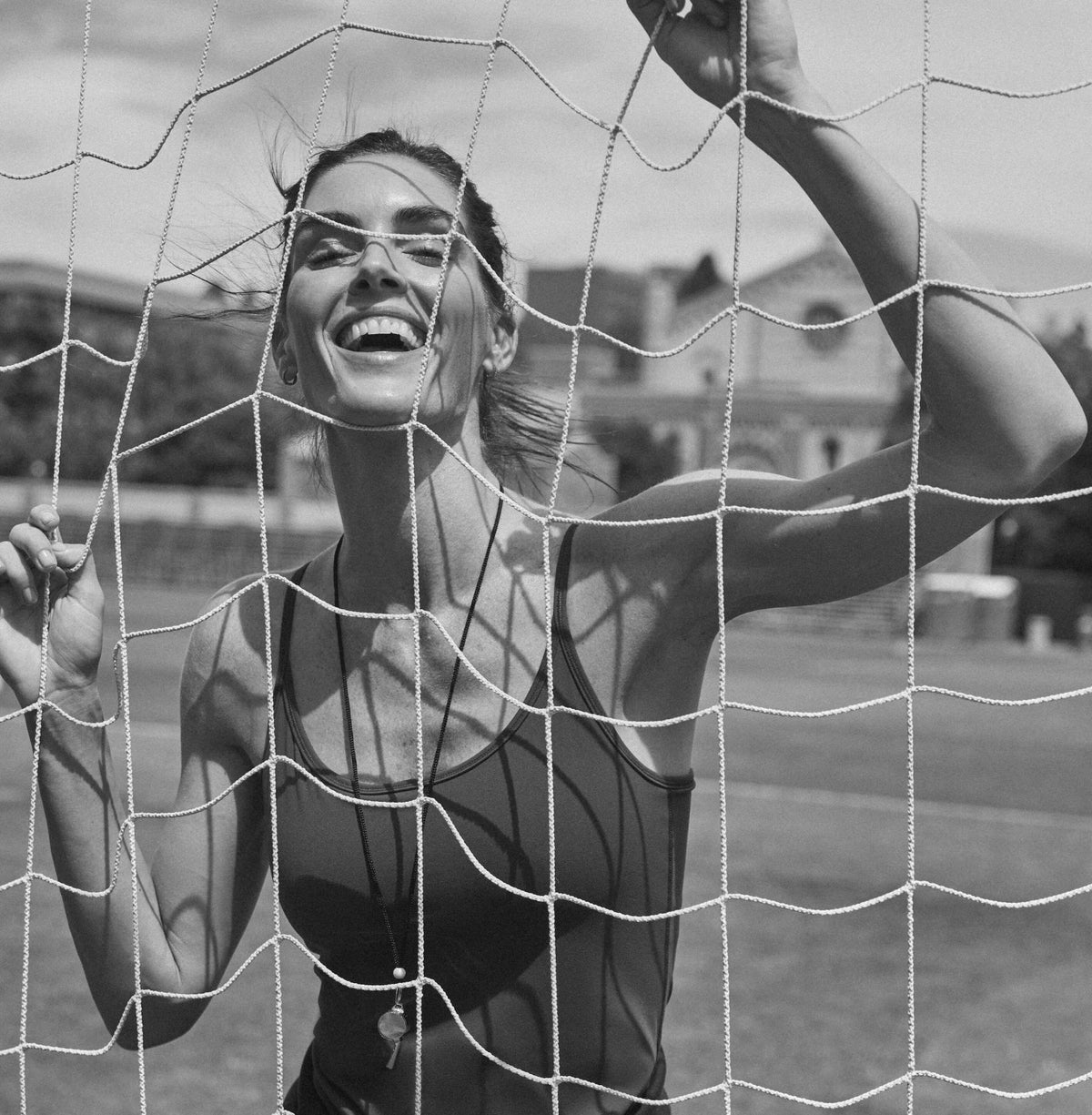 A woman in a sleeveless athletic top, smiling behind a sports net, exudes effortless fun and confidence. A whistle pendant adds a classic touch.