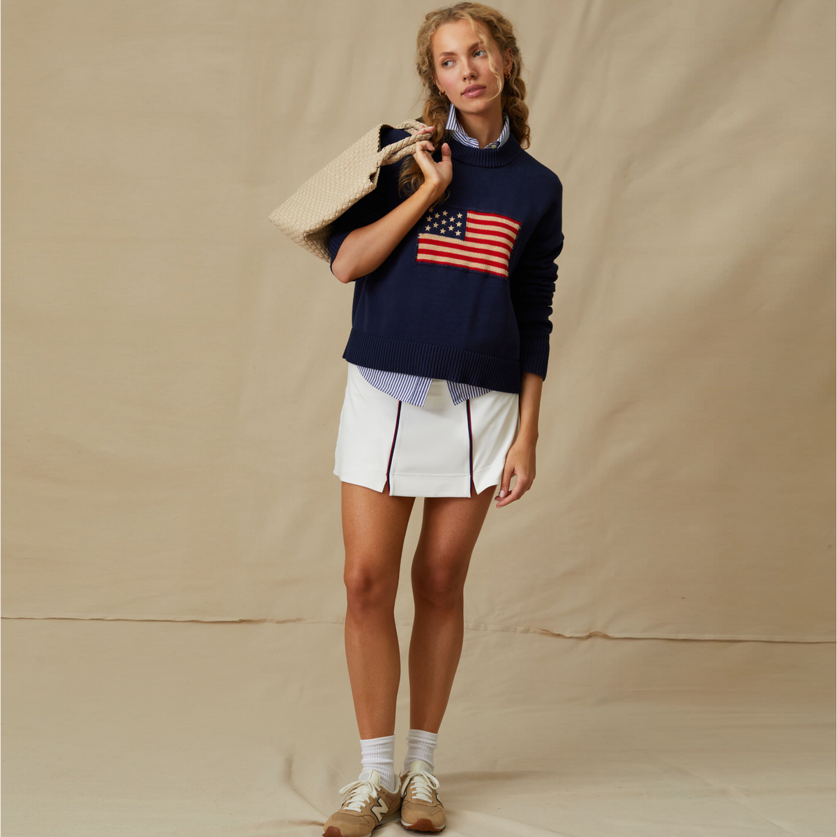 Young woman in a classic navy knit sweater with American flag, white skirt, and tan sneakers, carrying a woven tote, exuding effortless, high-end style.