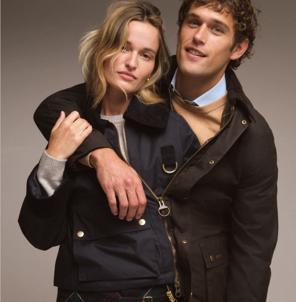 A man and woman in classic, high-end outerwear; navy jacket with gold accents, and waxed-cotton jacket, embodying effortless, fun style.