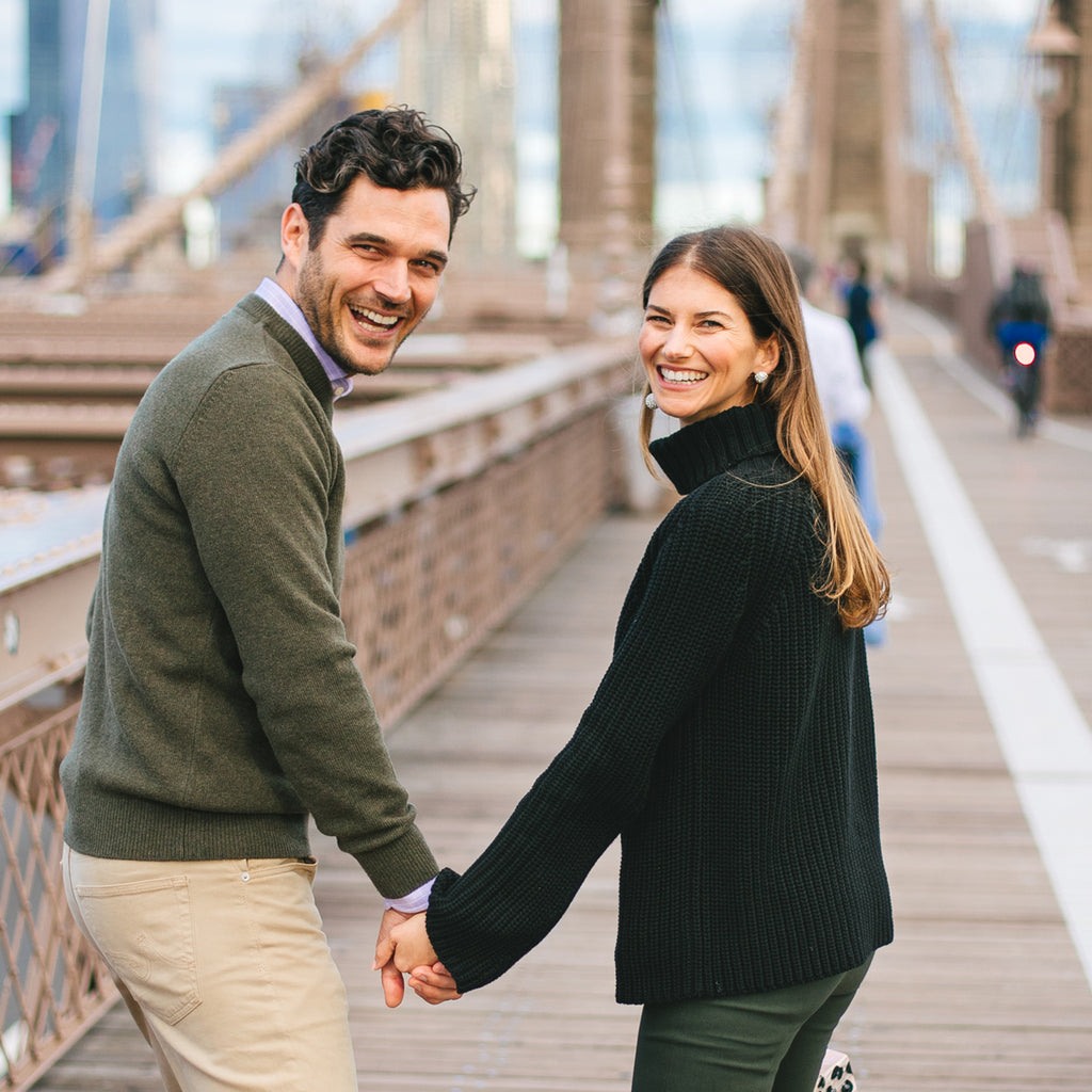 A man and woman in classic, high-end attire walk joyfully on a suspension bridge, embodying effortless style and fun, with a city skyline behind them.