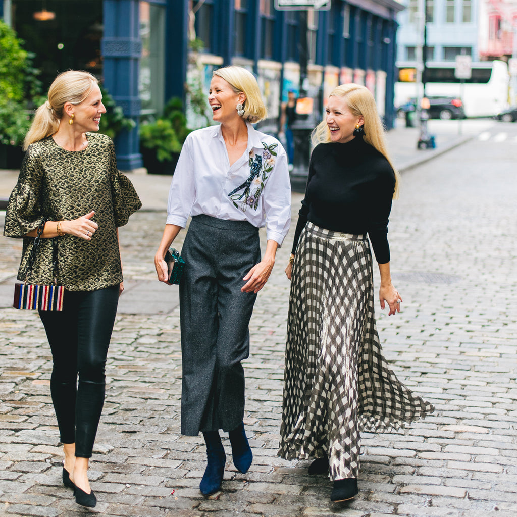 Three women in classic, fashion-forward outfits stroll a cobblestone street, exuding effortless style and fun in high-end Tuckernuck-inspired ensembles.