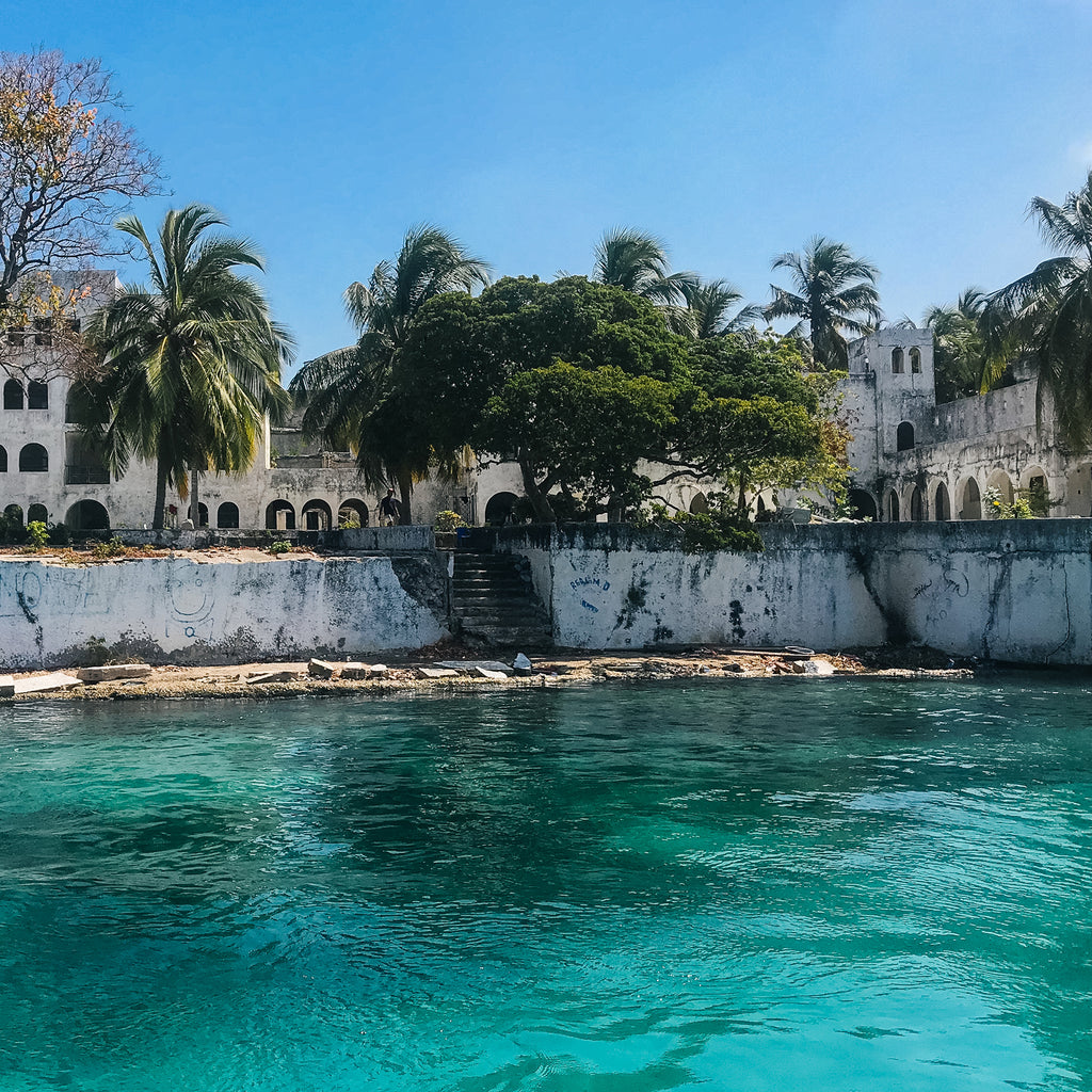 Tropical waterfront with classic colonial architecture, effortless charm, and fun, high-end vibes; arched doorways, palm trees, and emerald-blue water under a clear sky.