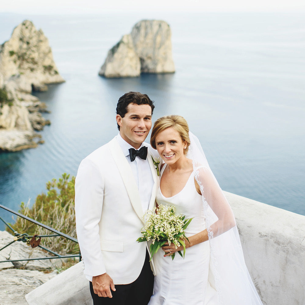 Newlywed couple in classic attire on seaside terrace; groom in white dinner jacket, bride in lace-edged veil, embodying effortless elegance and high-end style.
