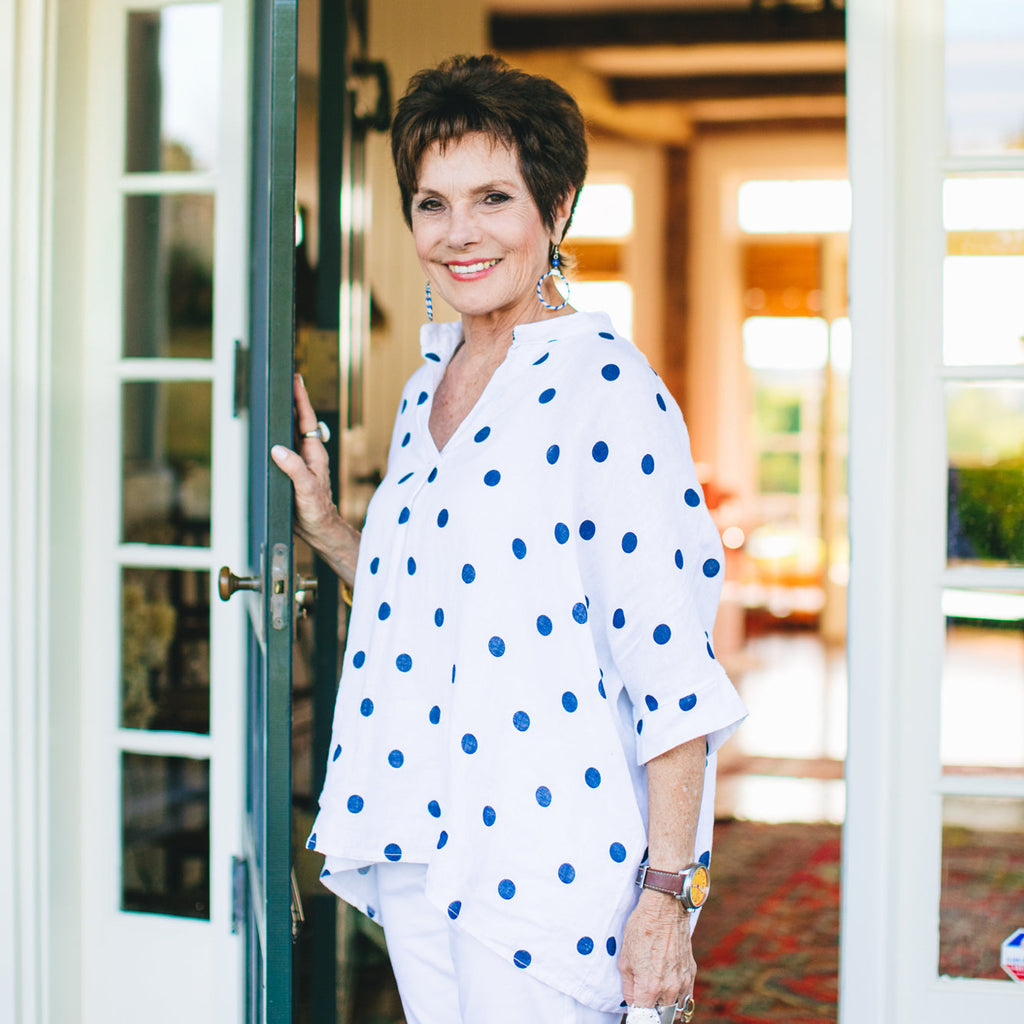 An older woman in a classic, effortless polka dot blouse stands smiling in a warmly lit doorway, epitomizing fun, high-end style.
