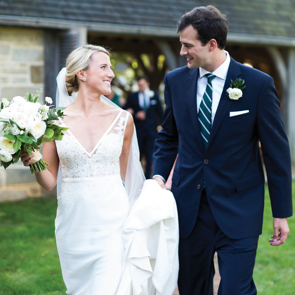 Newlyweds walk outdoors; bride in a classic lace gown, groom in a dark suit with boutonnière, embodying effortless elegance and high-end style.