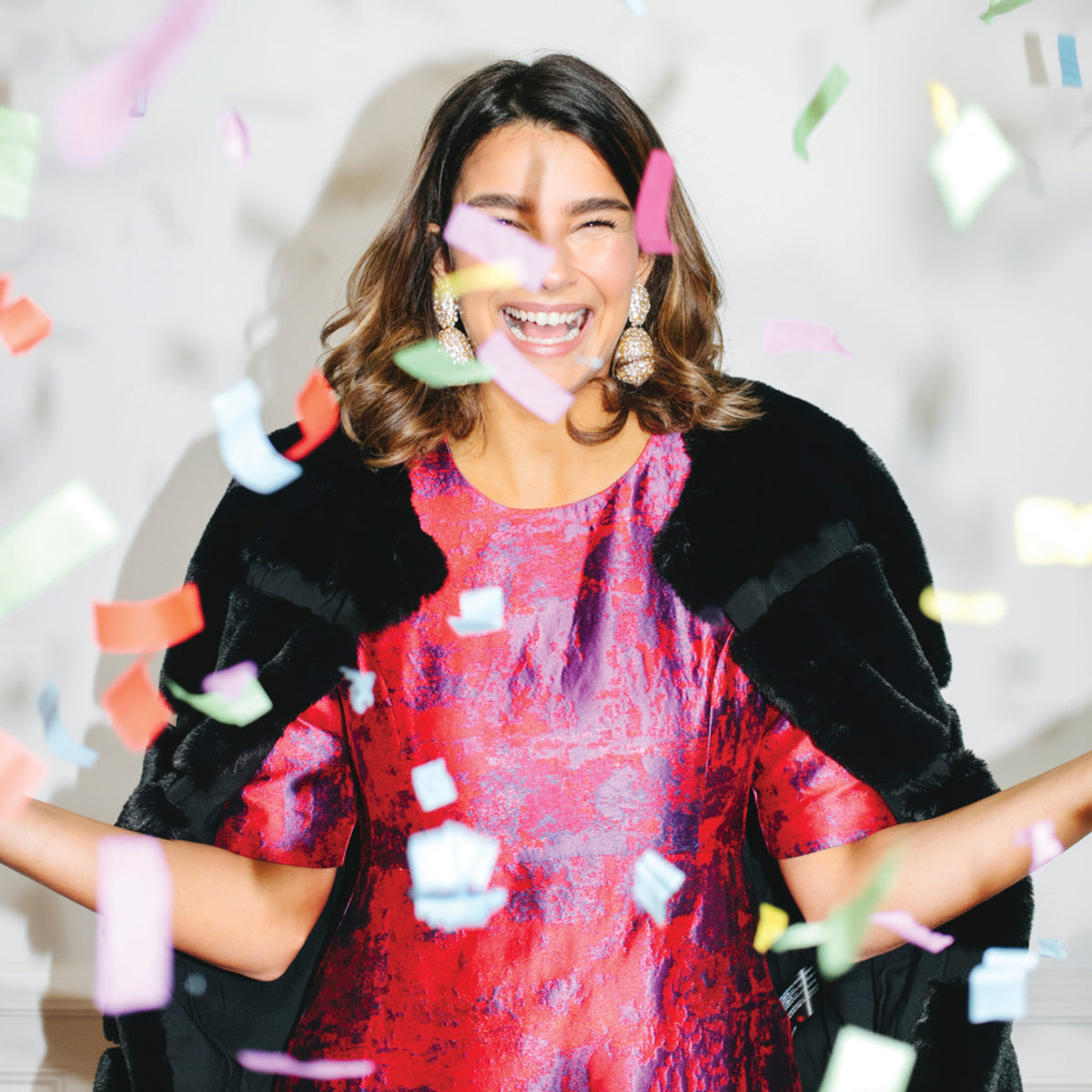 Woman celebrating in a shiny red dress with black fur-trimmed topper, statement earrings, and confetti falling around her, embodying effortless, high-end fun.