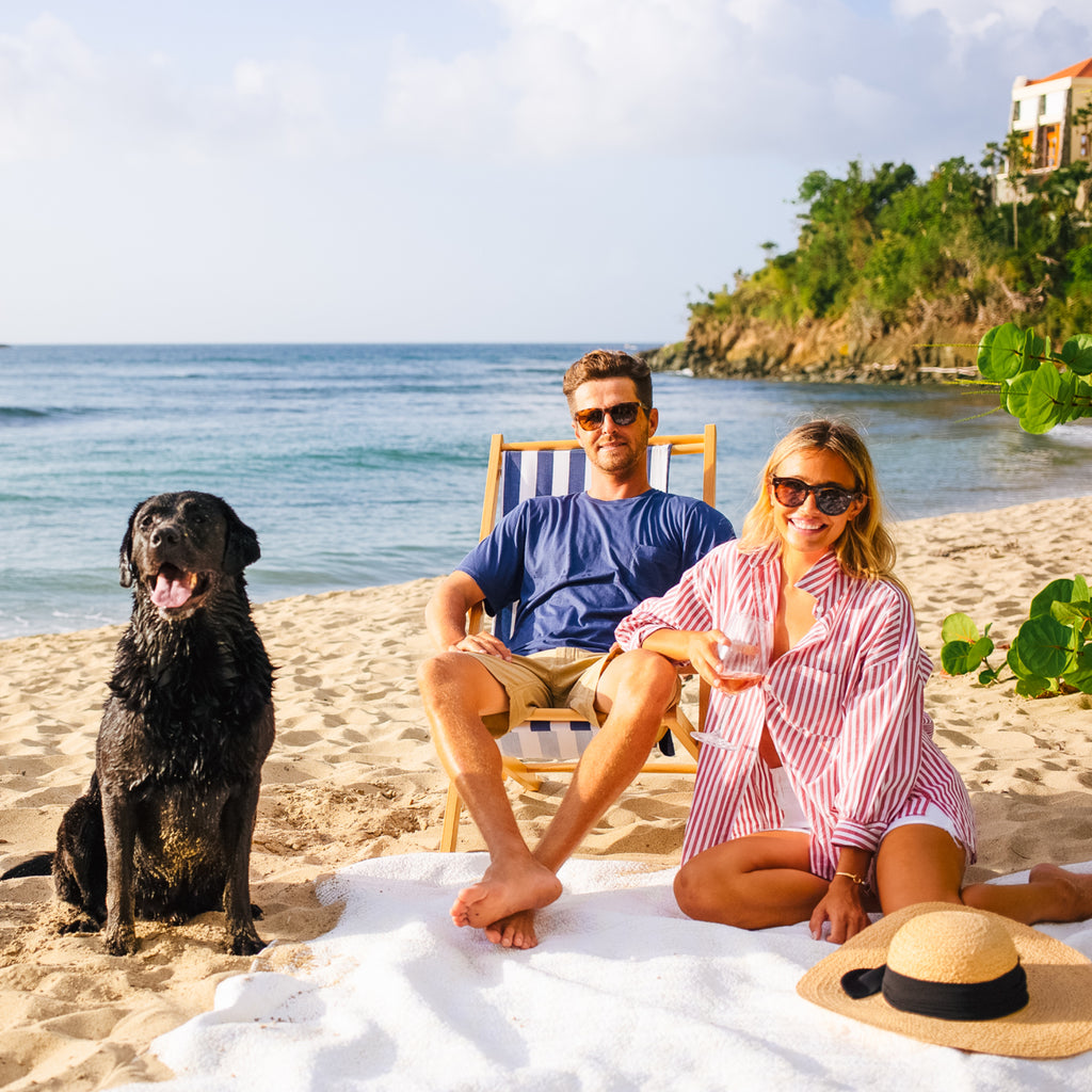 Sunlit beach scene with a man, woman, and dog; enjoying effortless, high-end relaxation. Classic summer attire and fun beach vibes reflect Tuckernuck's style.