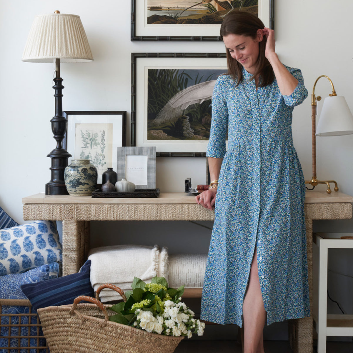 A woman in a classic blue floral dress stands by a woven-top console with lamps, prints, and vases, exuding effortless, high-end style.