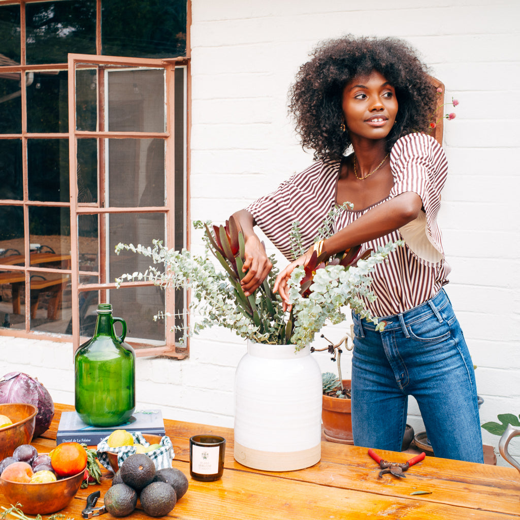 A young woman effortlessly arranges foliage in a classic ceramic vase surrounded by high-end fruits, gardening tools, and a cookbook on a stylish table.