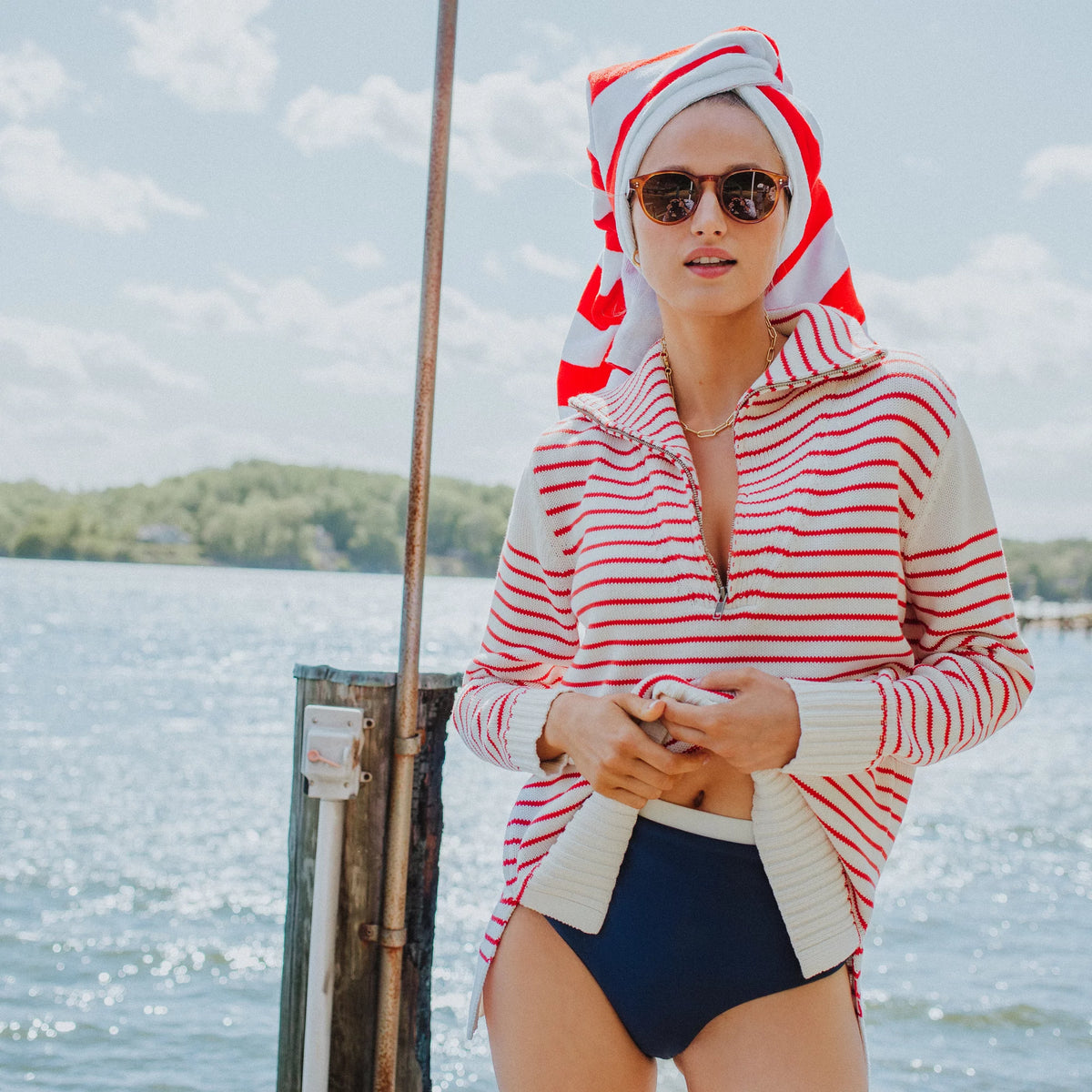 Young woman in classic red-and-white striped knit pullover, navy swim bottoms, round sunglasses; effortlessly styled on a dock, embodying fun and high-end leisure.