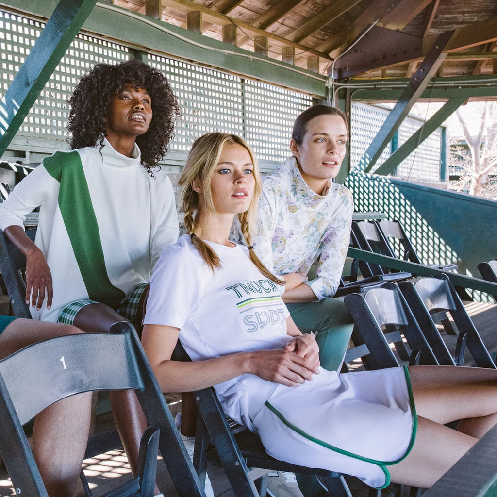 Three women in classic, tennis-inspired outfits sit on bleacher seats under a pavilion, embodying effortless, high-end style while enjoying a fun outdoor event.