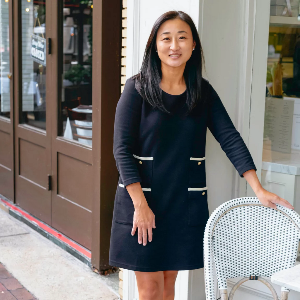 A woman in a classic, long-sleeved black dress stands outside a closed café, exuding an effortless, high-end style with a fun, urban vibe.
