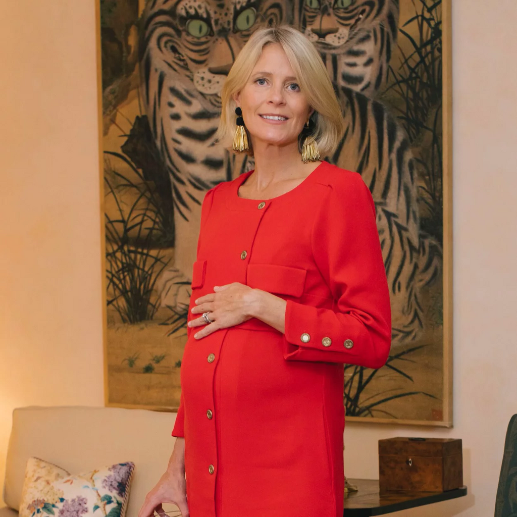 A woman in a classic red button-front coat, effortlessly accessorized, stands indoors, highlighting her pregnant belly. High-end decor and art enhance the scene.