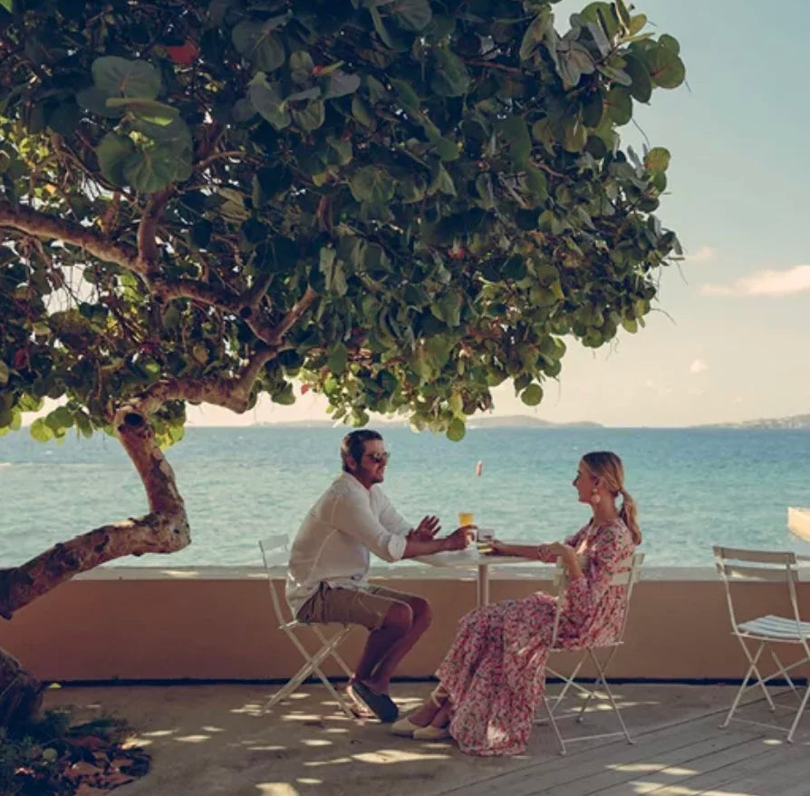 Man and woman enjoy drinks at a classic, white folding table on a terrace, under a tree, overlooking a calm sea, embodying effortless fun.