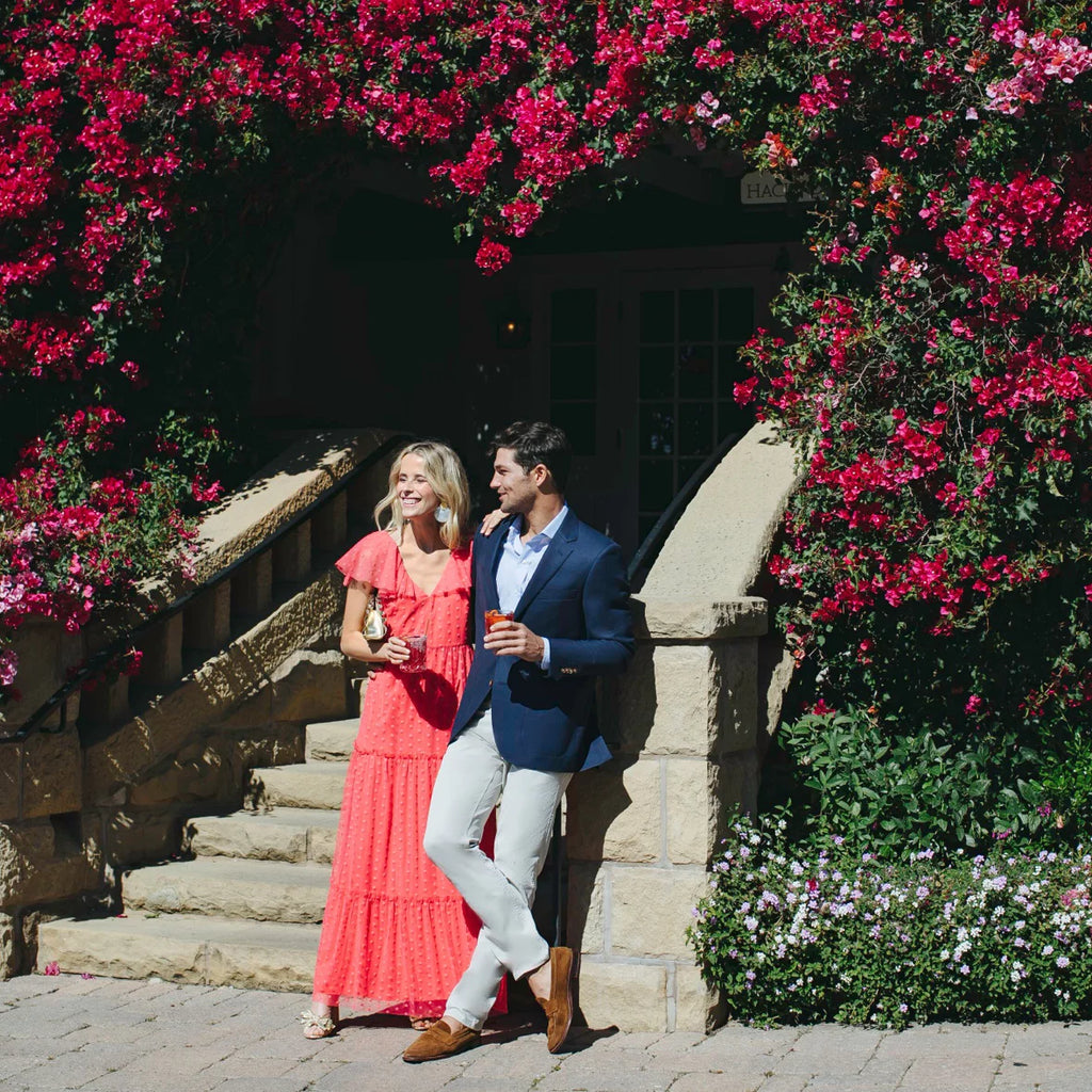 A couple exudes effortless elegance; she in a coral-red maxi dress, he in a navy blazer, amidst vibrant bougainvillea, embodying classic, high-end style.