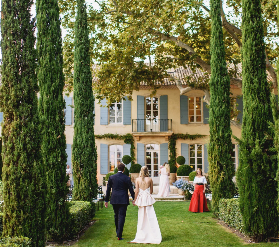 Bride in strapless gown and groom in dark suit stroll past cypress-lined path towards villa; classic, effortless outdoor wedding evokes fun, high-end elegance.