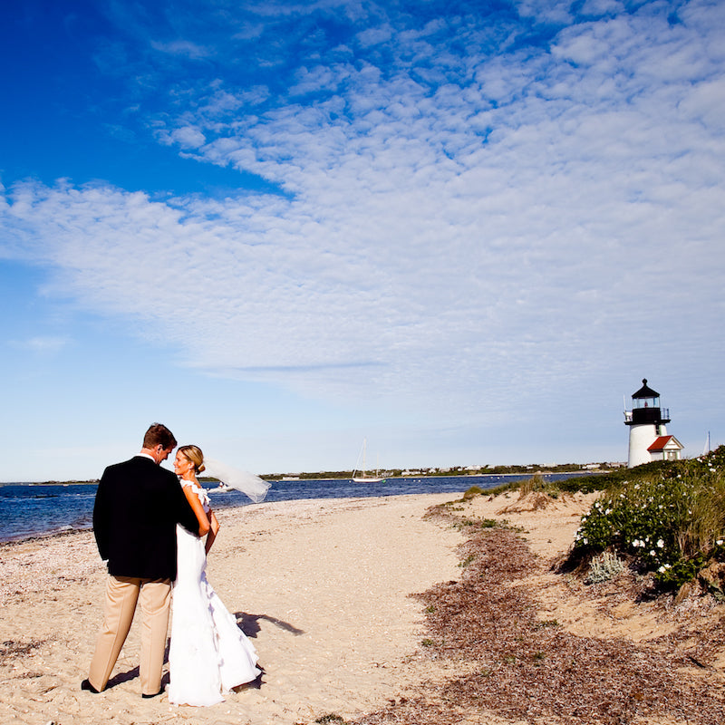 Bride and groom on a sandy beach near a classic lighthouse, embodying an effortless, high-end coastal vibe with fun sailboats in the background.