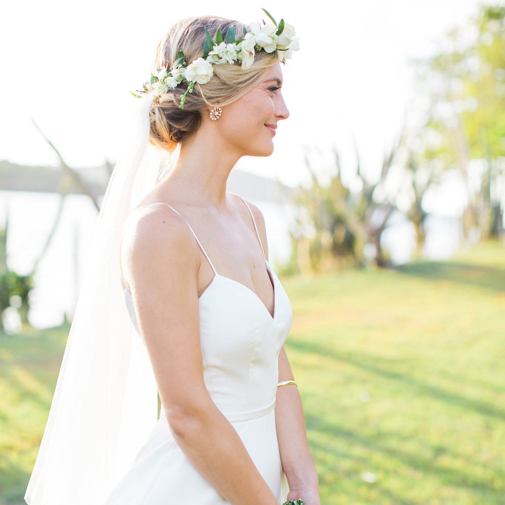 Bride in profile wearing a classic, effortless white gown with floral crown and veil, outdoors near water. Perfect for a high-end, fun wedding.