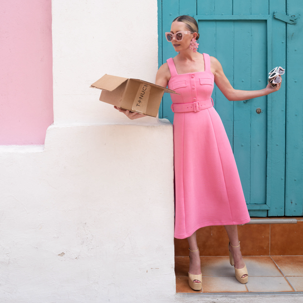 A woman in a classic pink midi dress holds T-NUCH box and sunglasses, exuding effortless, fun style with high-end accessories, embodying Tuckernuck's cool confidence.