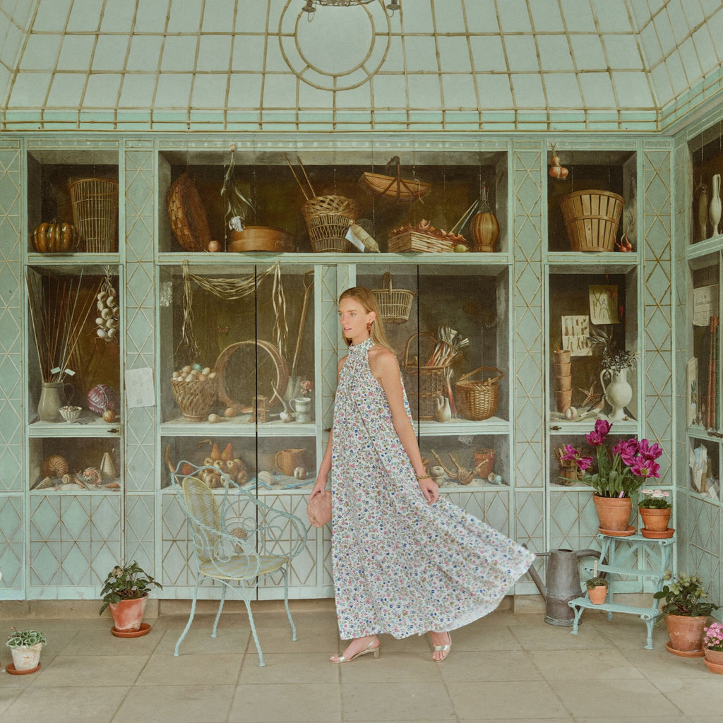 A young woman in a classic floral maxi dress strides through a light-filled space with garden curios, embodying effortless, high-end style and fun.