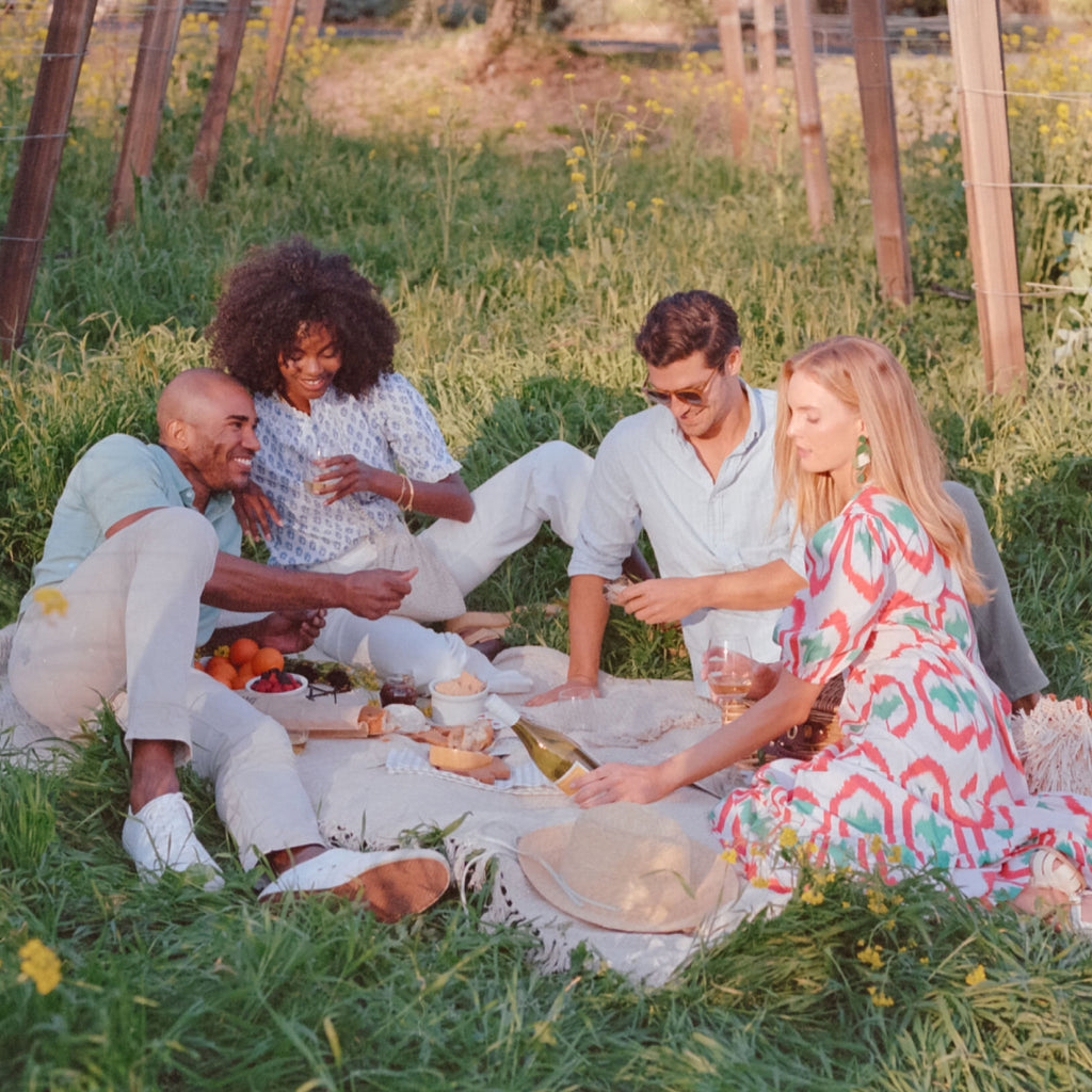 Four friends enjoy an effortless, classic picnic in a vineyard, sharing wine and gourmet snacks, embodying Tuckernuck's fun and high-end lifestyle.