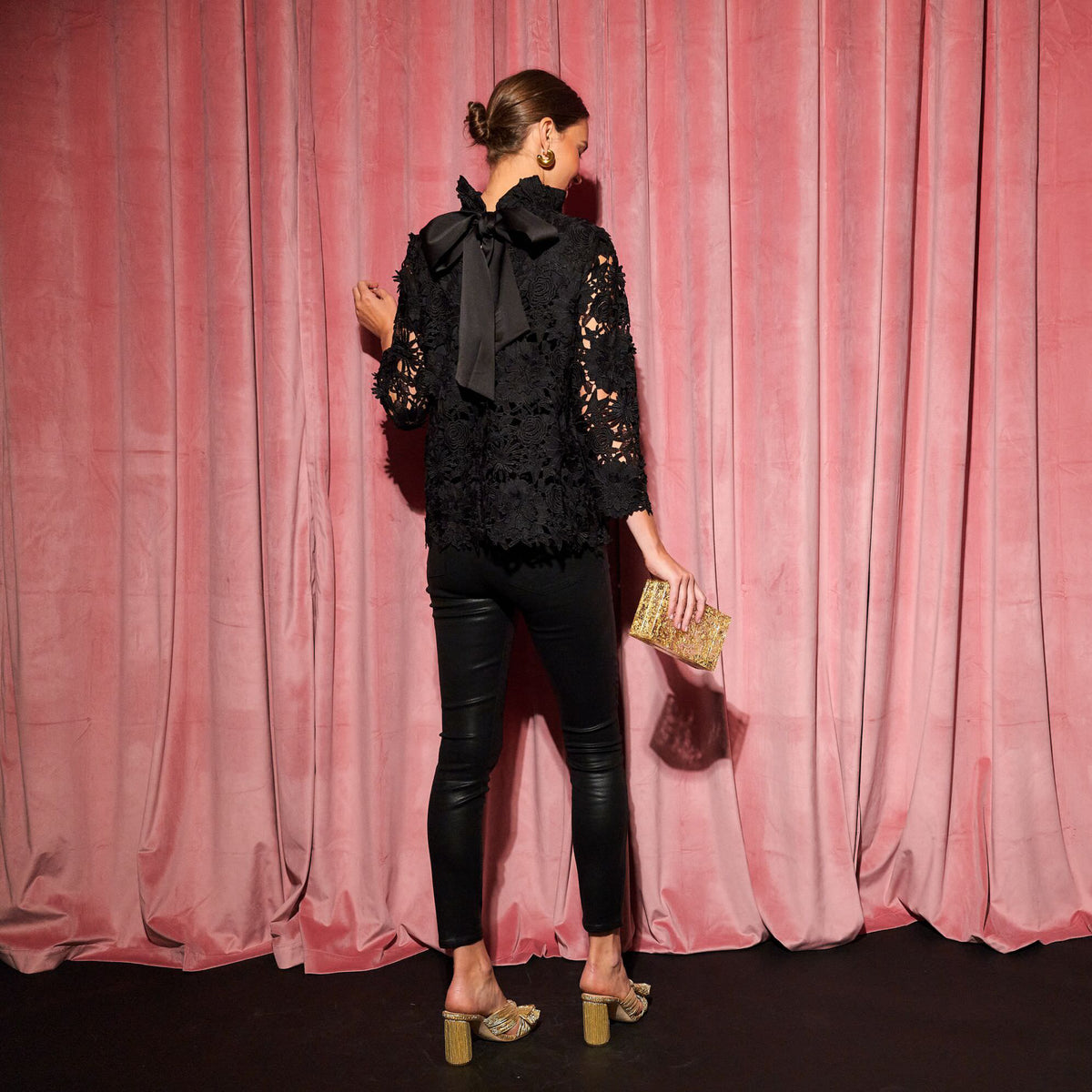 Person wearing a classic, all-black ensemble with floral-lace top and leather-look pants, holding a gold clutch, embodying effortless, high-end style against a velvet backdrop.