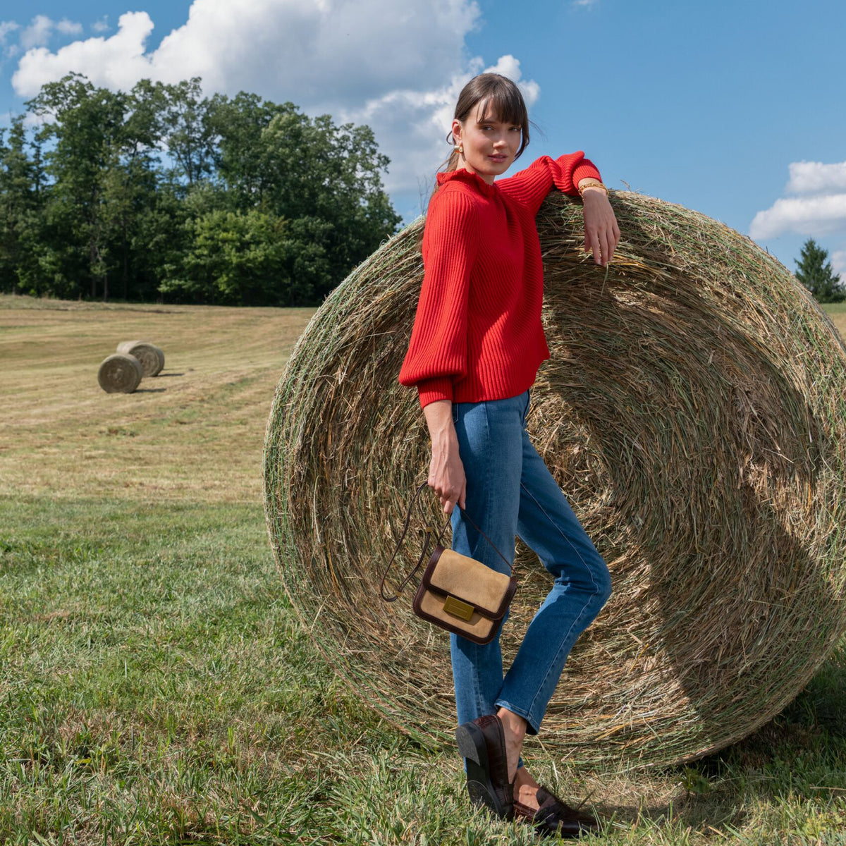 Young woman in a red sweater, blue jeans, and loafers, leaning on a hay bale with a tan shoulder bag; embodies classic, effortless style.