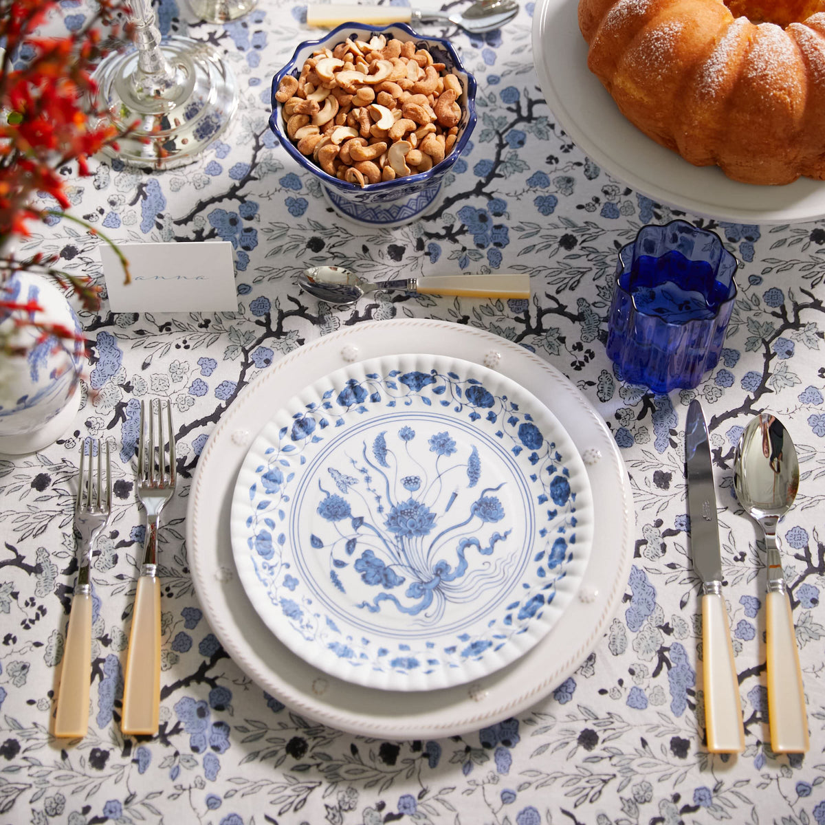 Overhead view of a classic, high-end blue-and-white floral table setting with porcelain plates, cutlery, cake, cashews, tumbler, and vibrant floral accents. Effortlessly fun.