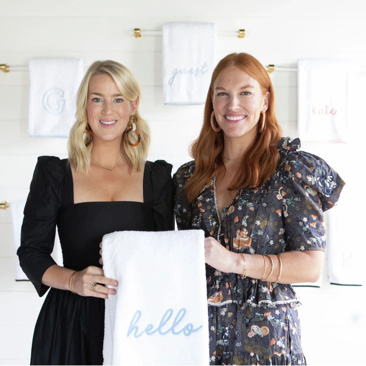 Two women display a classic, high-end white towel embroidered with hello, surrounded by effortlessly fun personalized bath linens on brass towel bars.