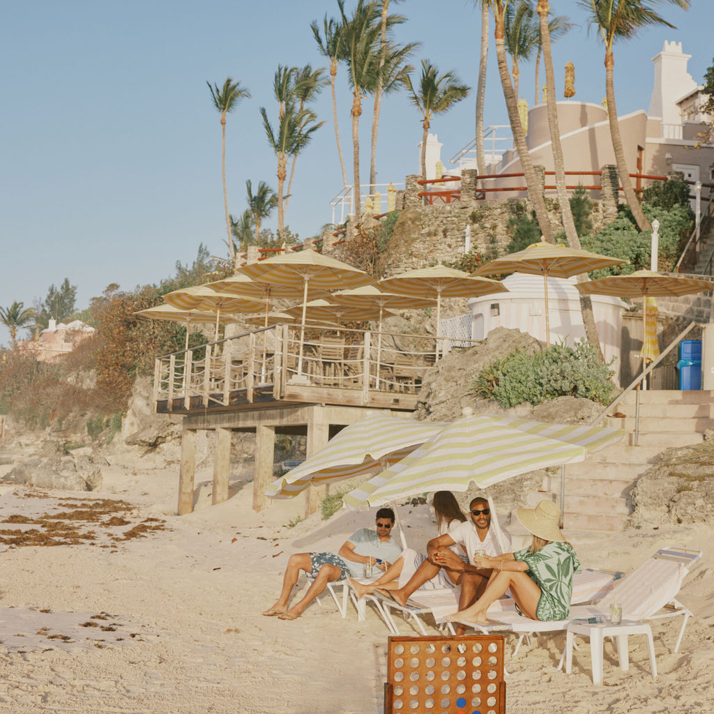 Friends lounging under classic yellow-and-white umbrellas on a beach, enjoying drinks, and playing Connect-Four; an effortless, fun, high-end seaside experience.