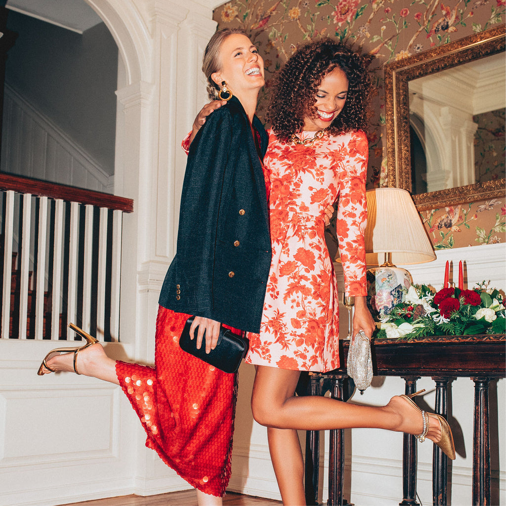 Two women in classic, festive attire showcasing effortless style and fun, dressed in high-end red ensembles with gold heels, embodying Tuckernuck's elegant spirit.