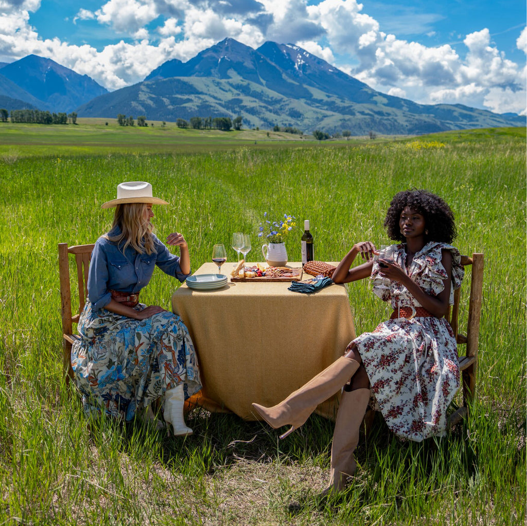 Two women enjoy a classic picnic in a meadow, featuring wine, charcuterie, and effortless style with floral attire, embodying high-end, fun elegance.