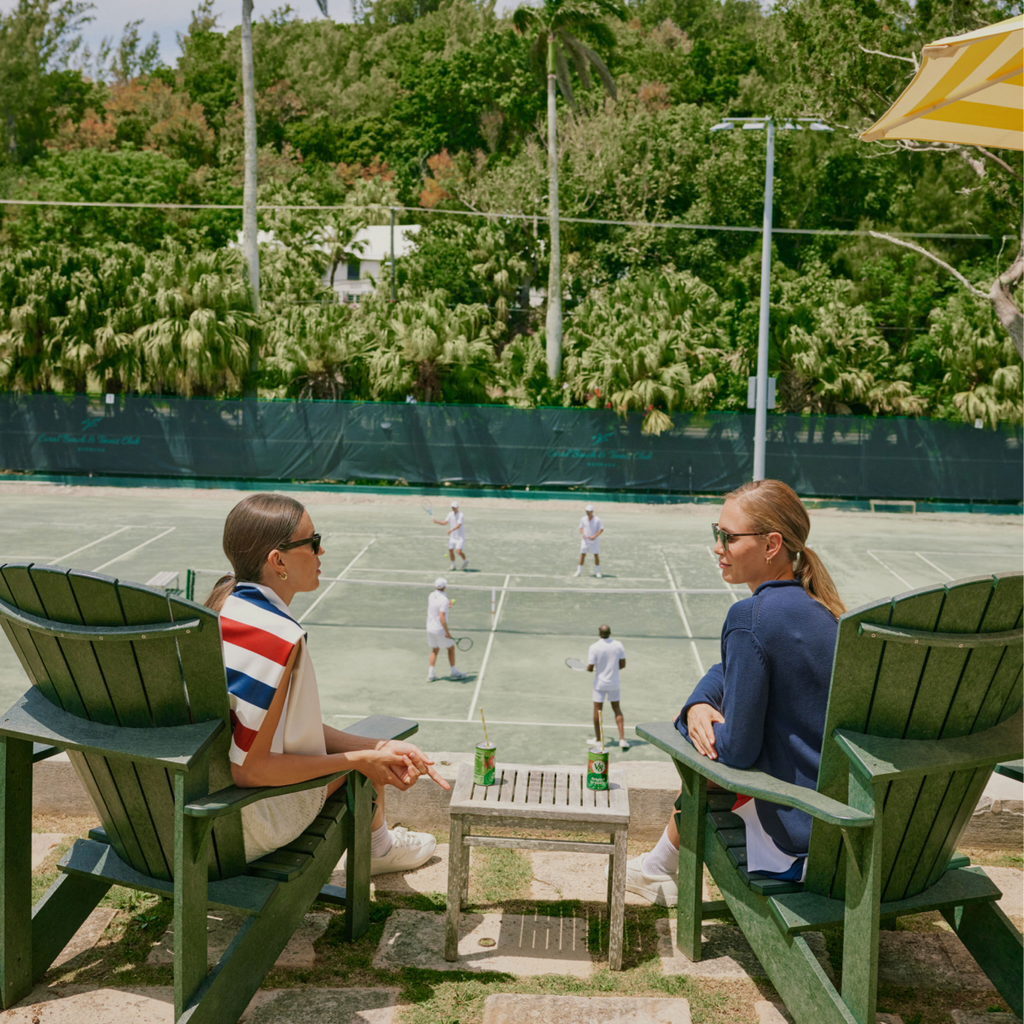 Two women in classic Adirondack chairs watch a doubles tennis match, enjoying V8 +Energy, embodying effortless, high-end leisure at a palm-lined club.