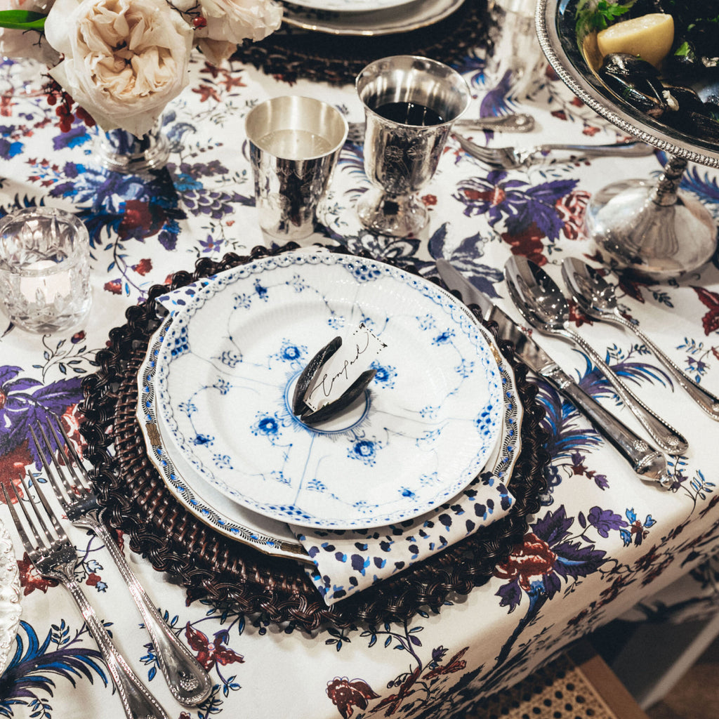 Elegantly set dining table features a classic floral tablecloth, porcelain plates, polished silverware, metal vessels, and a fun shell place-card holder for an effortless, high-end vibe.