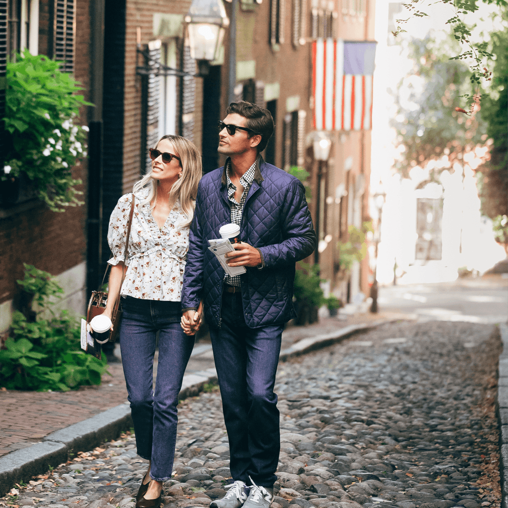 A casually dressed couple strolls a cobblestone street, exuding effortless style with sunglasses and coffee, embodying Tuckernuck's classic, fun, high-end vibe.