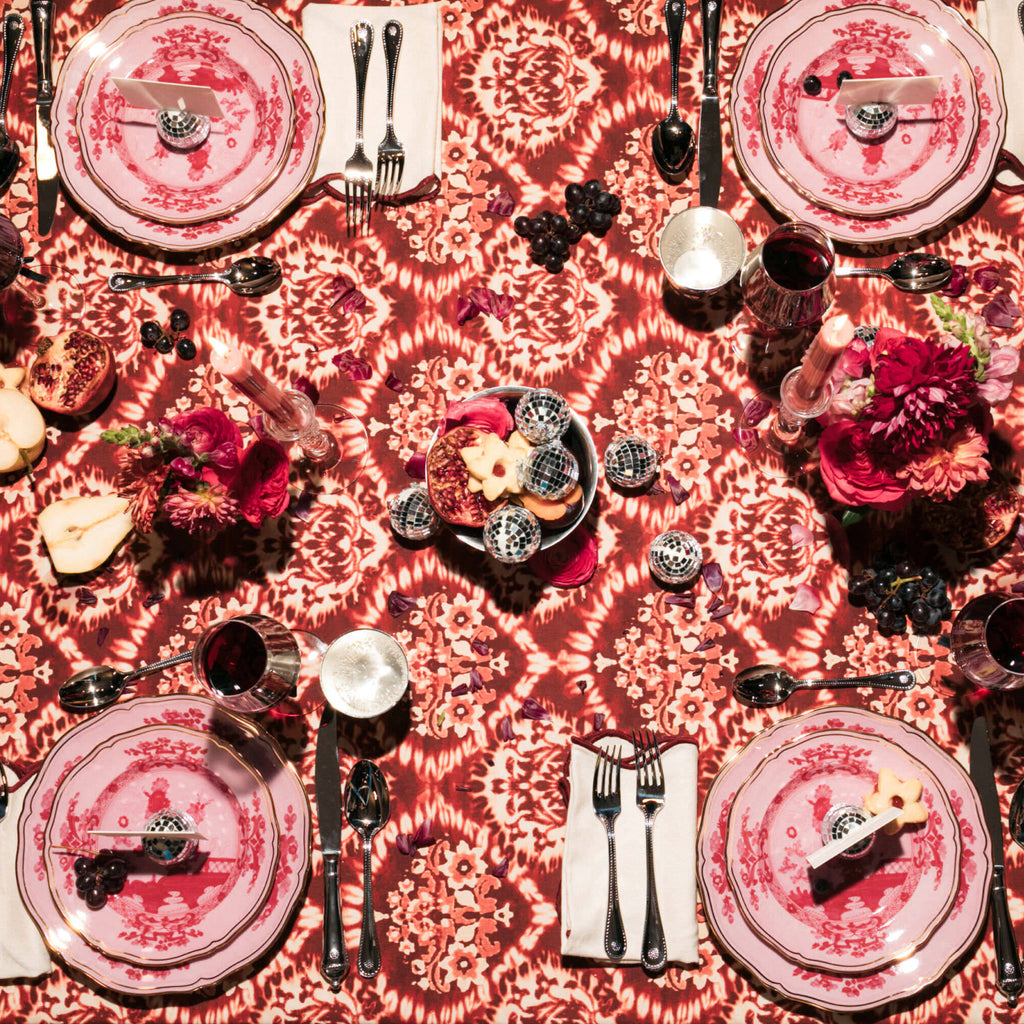 A classic, fun dinner table with effortless high-end flair: vintage plates, polished silverware, disco-ball ornaments, fruit, cookies, candles, and red-pink floral arrangements.