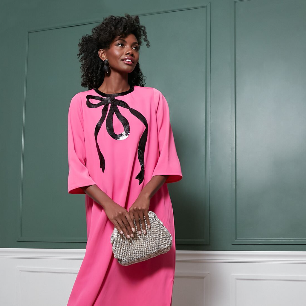 Woman in a hot-pink dress with a black sequin bow, holding a silvery beaded clutch; an effortless, fun, and high-end look.