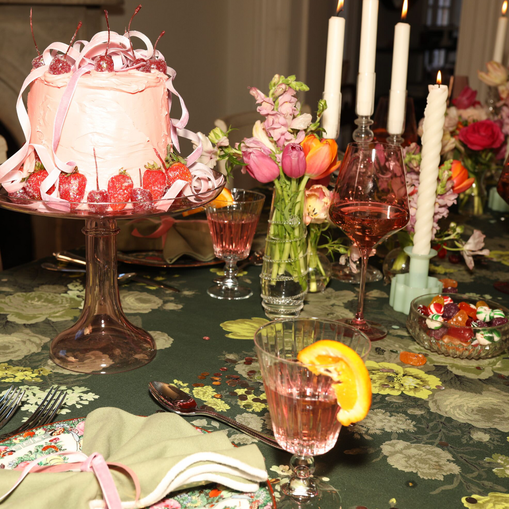Elegant table with a classic pink frosted cake, sugared berries, floral prints, rosé drinks, vibrant flowers, and glowing candles, creating an effortless, high-end celebration.