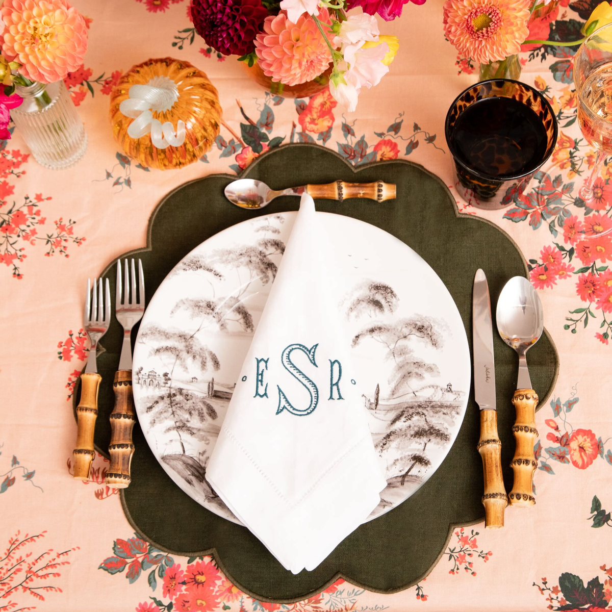 Elegant place setting on floral tablecloth with monogrammed napkin, bamboo cutlery, china plate, amber glass, and floral accents, evoking classic, effortless, high-end style.