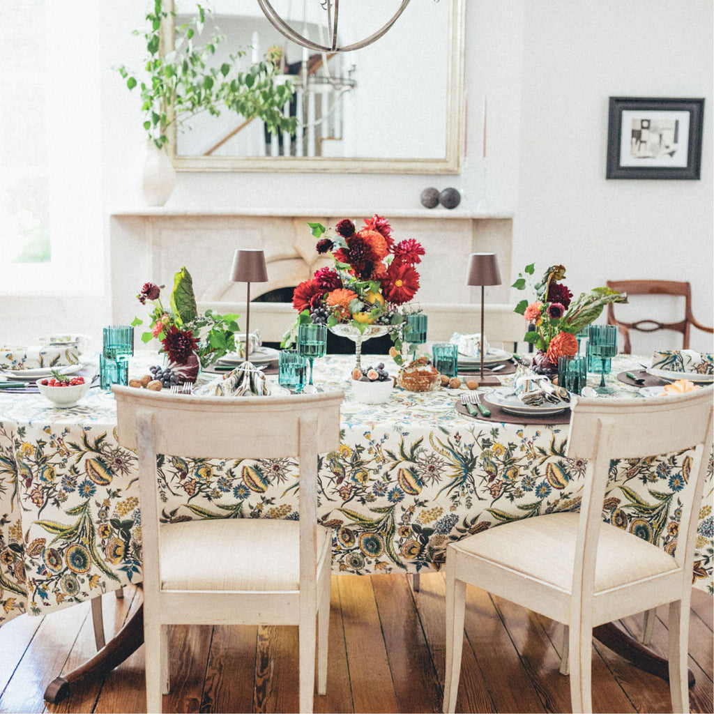 Bright dining room with a classic, floral table setting featuring emerald tumblers, vibrant centerpiece, and pale wooden chairs, evoking an effortless, high-end dining experience.
